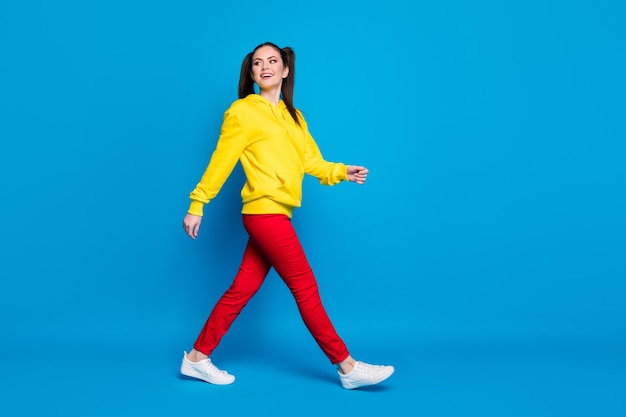 Full length profile photo of cheerful lady two tails good mood walk street see friends meeting wear casual yellow hoodie pullover red pants sneakers isolated bright blue color background