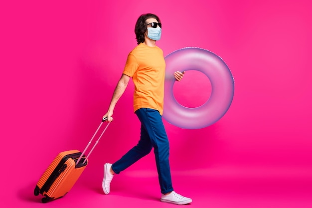 Full length profile photo of guy go hold case rubber circle wear t-shirt jeans medical mask isolated pink color background