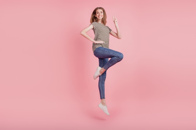 Full length profile side photo of young woman happy positive smile show peace cool v-sign jump up isolated over pink color background