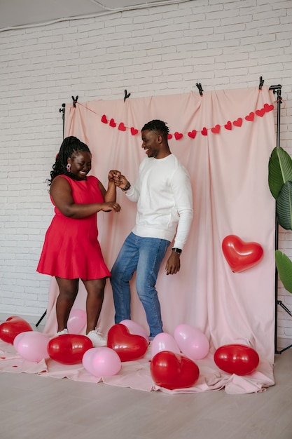 Full length of romantic young African couple dancing on pink background with heart shaped balloons