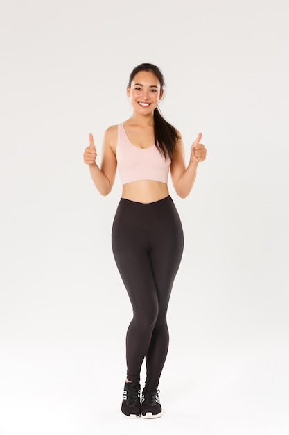 Full length of satisfied smiling, cute asian fitness girl, sportswoman in active wear showing thumbs-up and smiling pleased, proud female athelte gain daily workout goal, white background.