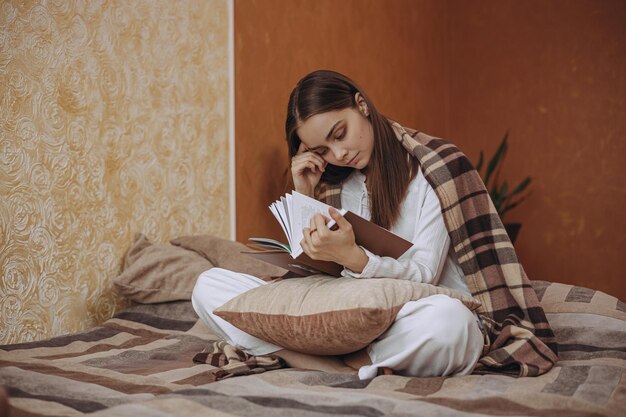 Full length of serene woman reading book and relaxing in warm plaid on bed in cozy room at home