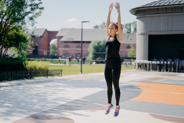 Full length shot of active young woman jumps high keeps arms raised up breathes deeply wears black t shirt leggings and sportshoes poses outdoors on stadium motivates you for going in for sport