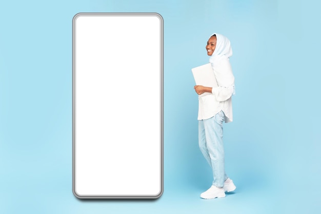 Full length shot of african american muslim woman holding laptop and walking near big cellphone with empty screen mockup