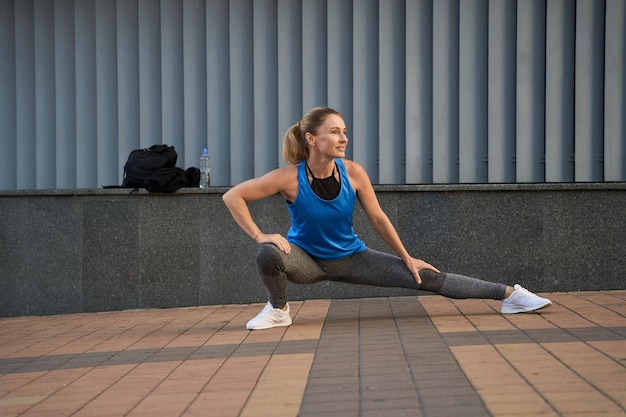 Full length shot of attractive sportive middle aged woman in sportswear looking away stretching her