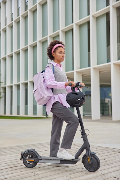 Full length shot of curly haired woman in casual clothes carries rucksack rides electric scooter in urban setting focused into distance uses eco transport at street near building during daytime