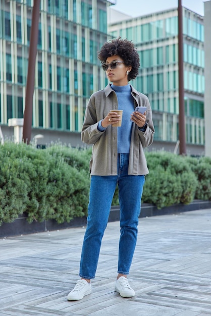 Full length shot of fashionable young woman wears jacket jeans white sneakers and sunglasses holds mobile phone and drinks coffee poses in urban setting. Modern lifestyle technology concept.