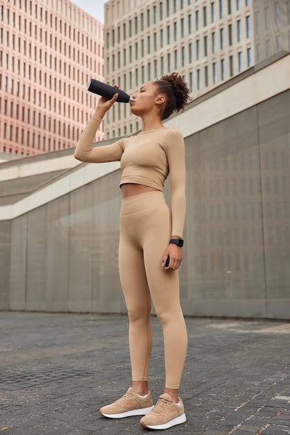 Full length shot of fit woman dressed in beige sportswear drinks fresh water after training leads sporty lifestyle has break during fitness workout exercises in open air People hydration and sport