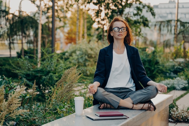 Full length shot of relaxed ginger woman meditates sits in lotus pose wears formal clothes keeps legs crossed breathes deeply spends free time in park surrounded by takeaway coffee tablet and notepad