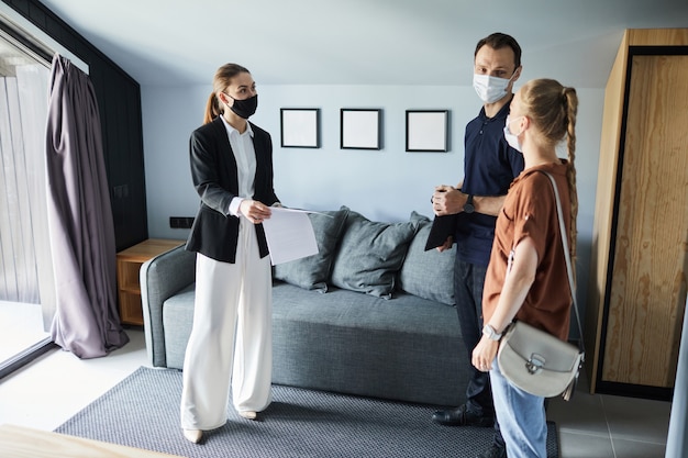 Full length side view portrait of female real estate agent wearing mask while signing contract with young couple buying new house, copy space