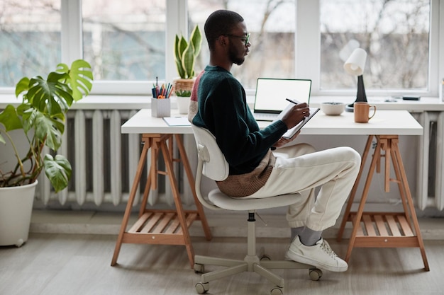 Full length side view portrait of young africanamerican man wearing glasses while working from home ...