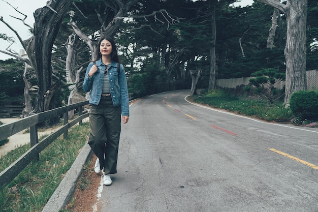 full length smiling asian girl backpacker walking alone on mountain highway in spring cloudy. vintage style of young lady on road near forest. happy elegant chinese woman on 17 mile drive california.