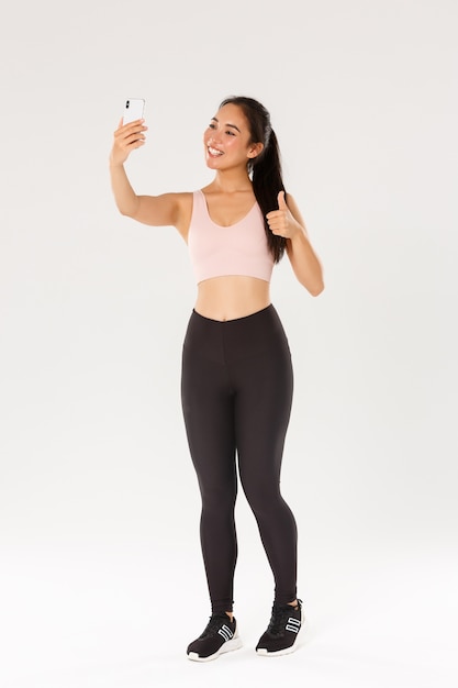 Full length of smiling female lifestyle blogger, asian fitness girl in sportswear taking selfie in gym, showing thumbs-up, demonstrate workout exercies for followers online, live stream from training.