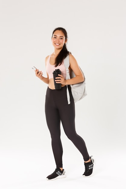 Full length of smiling healthy, slim asian girl going fitness training, female athelte carry backpack with workout equipment and water bottle, using mobile phone sports application, white background.