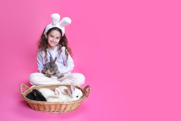 Full length of a smiling little girl with bunny ears straw basket with little rabbits isolated on a pink background