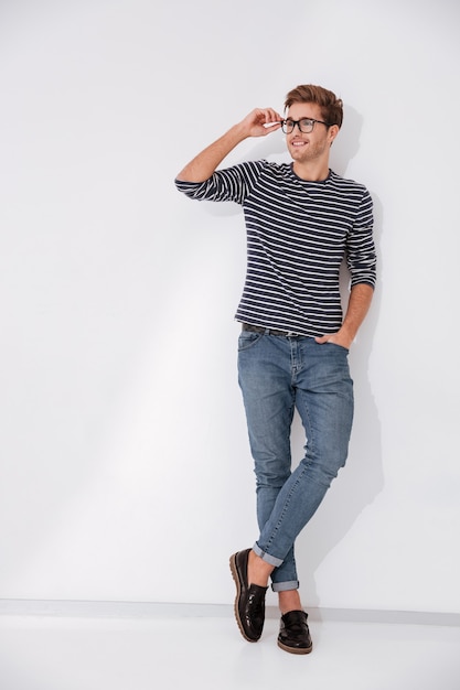 Full length smiling man in striped sweater posing in studio with arm in pocket and holding his eyeglasses. Isolated gray background