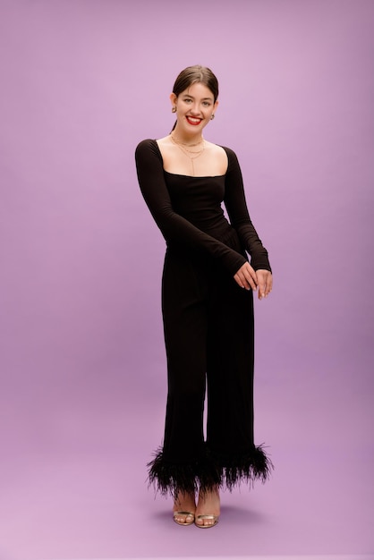 Full length smiling young caucasian brunette woman with red lips in black elegant suit on purple background