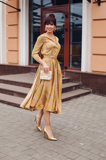 Full length stock photo of a gorgeous elegant lady in golden dress, high heels of golden color and crossbag. She is walking in the street smiling.