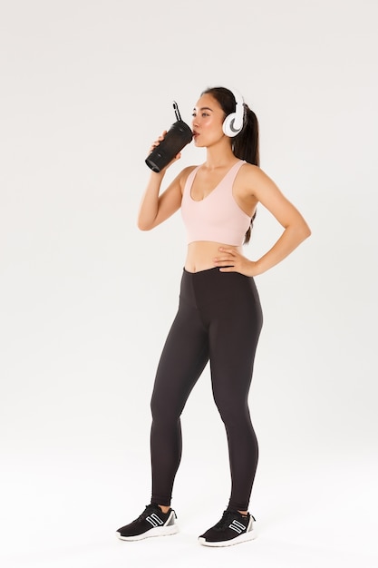 Full length of sweaty pleased female athelte drinking water from bottle while running, prepare to marathon. Fitness girl in sportswear, listening music headphones during workout 