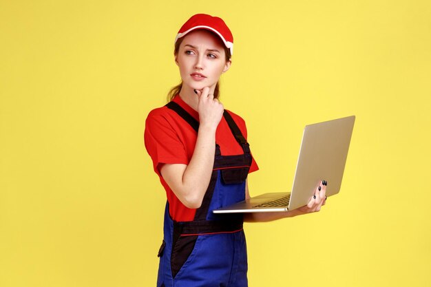 Full length thoughtful uncertain worker woman holding laptop, pondering decision, online order for repair services, wearing overalls and red cap. Indoor studio shot isolated on yellow background.