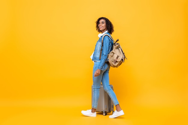 Full length travel portrait of amazed young African American woman backpacker walking with luggage on studio yellow studio background