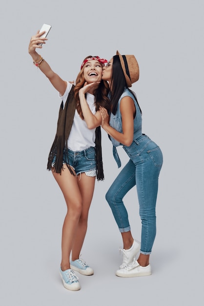 Full length of two attractive stylish young women taking selfie and smiling while standing against grey background