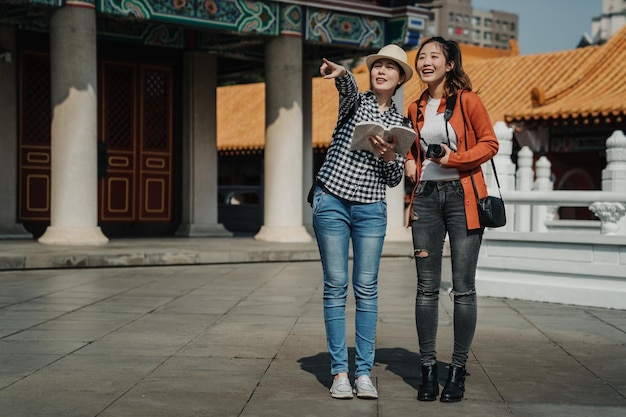 full length of two beautiful asian girlfriends walking on tour trip in beijing china. japanese ladies tourists standing on ground girl showing point finger chinese garden view in confucius temple.