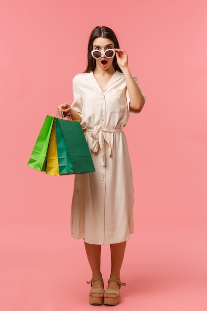 Full-length vertical portrait excited and speechless, amazed young brunette woman seeing something amazing and pretty, taking off glasses to look closer, open mouth fascinated, holding shopping bags