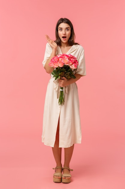 Full-length vertical portrait pretty surprised girl receive unexpected gift, got delivery holding flowers, gasping open mouth amazed and looking speechless with stunned expression, pink wall