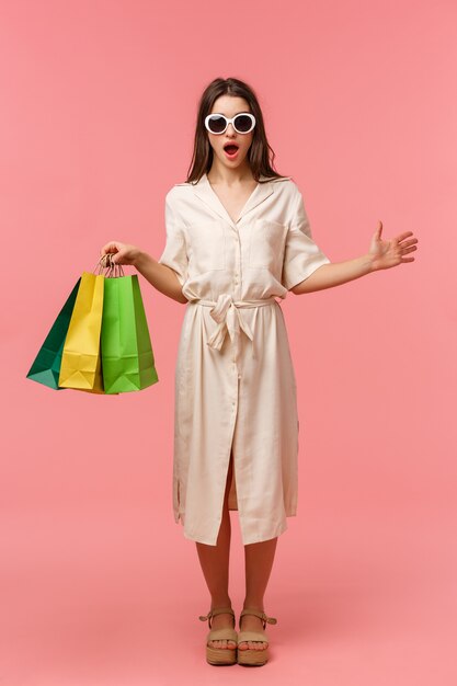 Full-length vertical portrait surprised and excited attractive feminine girl in dress and sunglasses, holding shopping bags, seeing someone in store, gasping amazed, standing pink wall