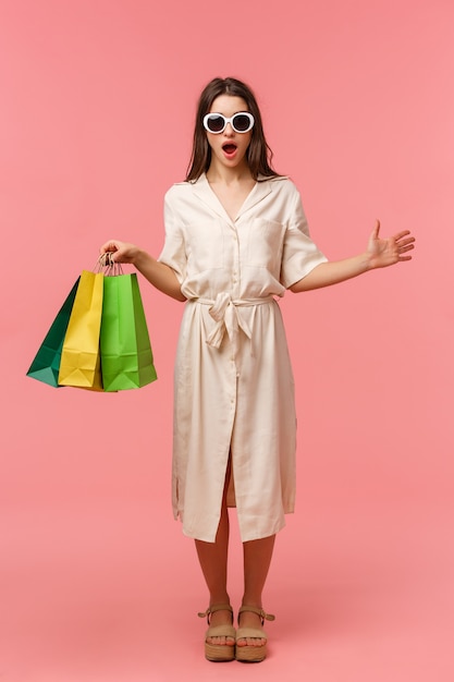 Full-length vertical portrait surprised and excited attractive feminine girl in dress and sunglasses, holding shopping bags, seeing someone in store, gasping amazed, standing pink 