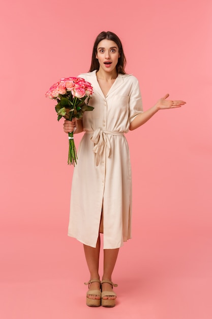 Full-length vertical portrait surprised see boyfriend with flowers, receive charming bouquet, spread hand sideways and shrugging surprised, look wondered and astonished, standing pink wall