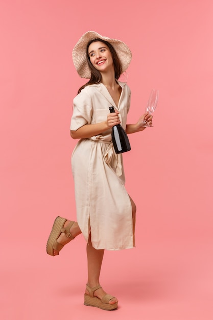 Full-length vertical shot woman on picnic, going out with girlfriend on fine sunny day in hat and dress, rushing with bottle champagne and glasses, smiling happily, standing pink wall