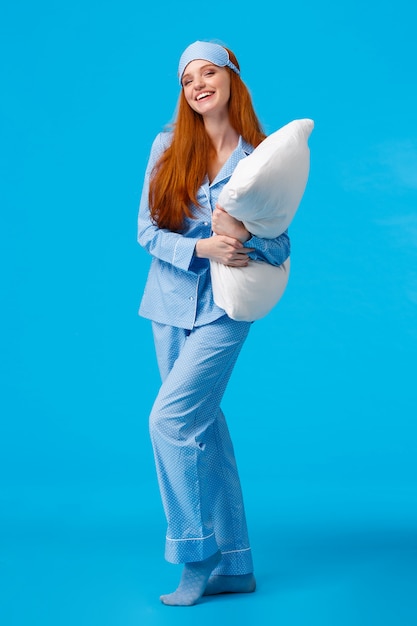 Photo full-length vertical studio cheerful lovely caucasian woman with ginger red long hair in nightwear, pyjama and sleep mask, hugging pillow ready sleep going bed at nigh and laughing, smiling