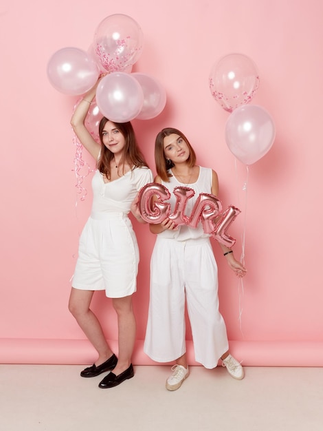 Full length view of smiling two girl dressed in a white holdings a balloons isolated pink background