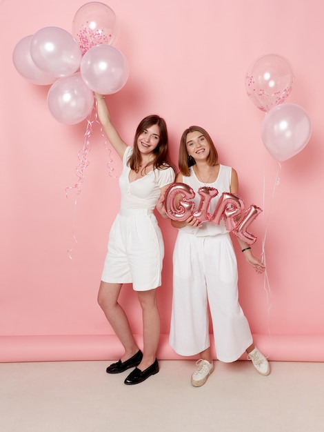 Full length view of smiling two girl dressed in a white holdings a balloons over pink background