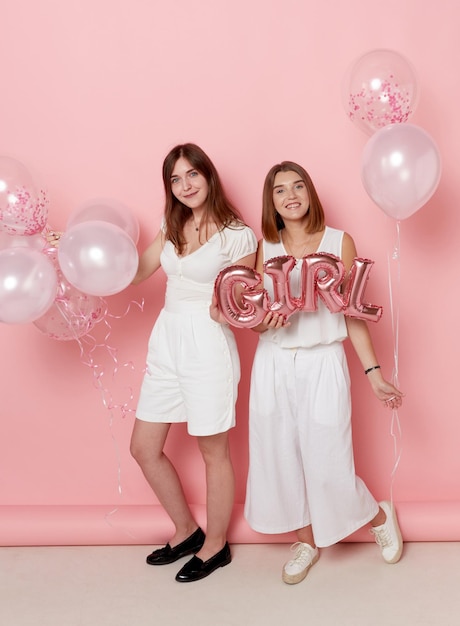 Full length view of a two of young friends dressed in a white holdings a balloons over pink background