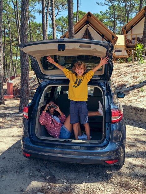 Full length of woman looking through binoculars and smiling boy standing in trunk of car parked near glamping huts placed in woods during summer trip