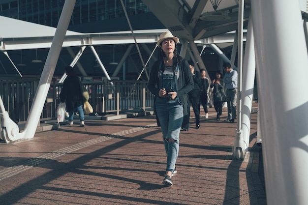 full length young asian woman tourist taking walk on footbridge while sightseeing in osaka city. beautiful girl holding camera relaxing on overpass with local people in background on sunny day.