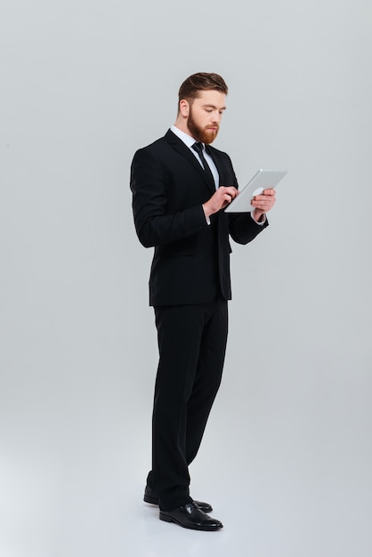 Full length young bearded business man in black suit standing sideways and writing message on tablet computer Isolated gray background