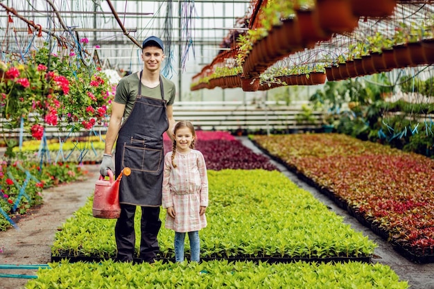 Full length of young happy worker and small girl standing at plant nursery and looking at camera