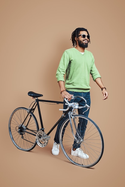 Full length of young man in casual clothing walking with bicycle while standing against brown wall