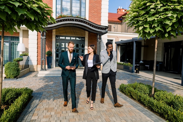 Full length of young three smiling multiracial businesspeople discussing about a meeting while walking outdoors in front of modern building, office or restaurant or hotel