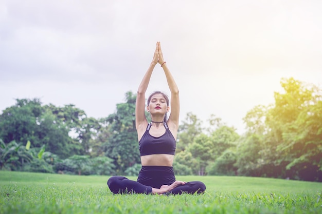 Full length of young woman with arms raised meditating while sitting on grassy field against sky in park