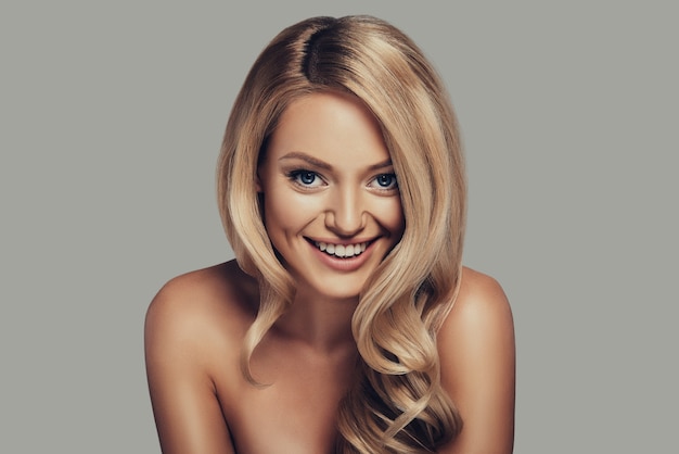 Full of life energy. Attractive young woman with long curly blond hair smiling and looking at camera while standing against grey background