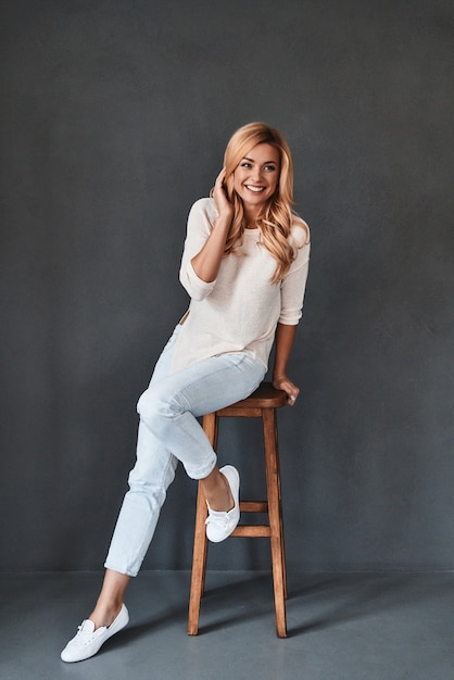 Full of life energy. Full length studio shot of attractive young woman looking at camera and smiling while leaning on stool against grey background