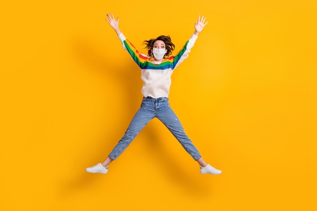 Full size photo of crazy lady jump like star rejoicing wear medical mask sweater jeans footwear isolated on yellow background