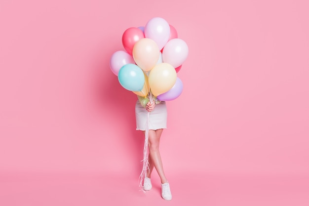 Full size photo of funny lady come birthday party hold many air balloons hiding face surprise visit wear casual green crop pullover jeans skirt shoes isolated pink pastel color background