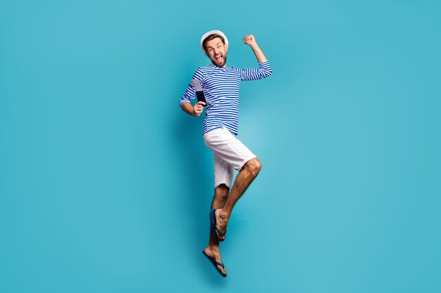 Full size profile photo of funky crazy guy tourist jump high celebrate vacation start finally got visa wear striped sailor shirt vest cap shorts flip flops isolated blue color