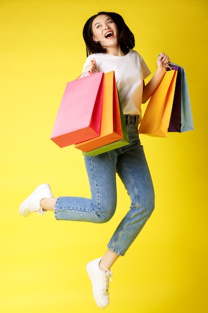 Full size shot of beautiful asian woman jumping with shopping bags and smiling happy, yellow background